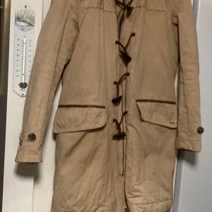Beige trenchcoat från Thomas Burberry - Säljer en klassisk beige trenchcoat från Thomas Burberry. Jackan har långa ärmar och knytning framtill. Den är tillverkad i 100% bomull och har dekorativa detaljer som knappar och en liten metallapplikation med TB-logga.