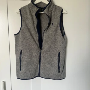 Grå väst från polo Ralph lauren  - Snygg grå väst från Ralph Lauren med dragkedja och två fickor framtill. Västen har en stilren design med mörkblå detaljer vid kanterna och en liten logotyp på bröstet. Storleken är XL för barn och ska symbolisera 18-20 år. Skulle säga att den passa S-M i vanlig storlek och materialet är fleece