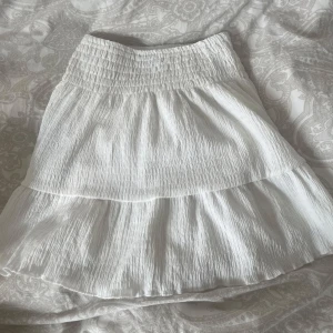 Vit kortkjol med volang - Säljer en super söt vit kortkjol med volangdetaljer!!💘Kjolen har en elastisk midja för en bekväm passform och är perfekt för varma sommardagar. Den är gjord i ett lätt och luftigt material som ger en fin rörelse. Passar ca 12-14 åringar.