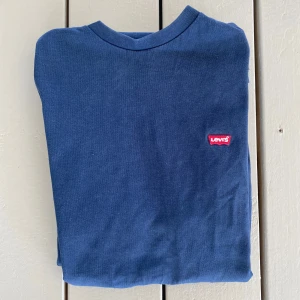 Blå t-shirt från Levi's - Säljer en klassisk blå t-shirt från Levi's med en liten röd logga på bröstet. T-shirten är kortärmad och tillverkad i mjuk bomull. Perfekt för en avslappnad stil. Priset är inte hugget i sten så hör av dig