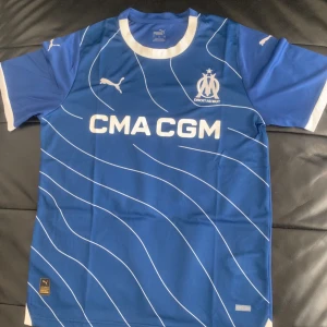 Marseille fotbollströja - Säljer en blå fotbollströja från Puma med vita detaljer och vågiga linjer. Tröjan har korta ärmar och klubbens logga på bröstet. Perfekt för fotbollsfans!