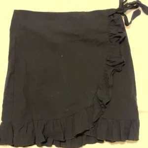 Svart omlottkjol med volang - Snygg svart omlottkjol med volangdetaljer och knytning i midjan. Perfekt för en stilren look. Kjolen har en kort längd och är lätt att matcha med olika toppar.