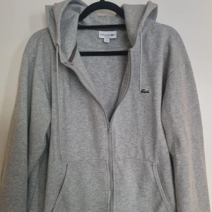 Grå hoodie från Lacoste - Grå hoodie från Lacoste med dragkedja och huva. Tröjan har långa ärmar och krokodilloggan på bröstet.