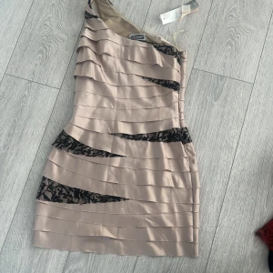 Champagnefärgad klänning från Lipsy - Elegant champagnefärgad klänning från Lipsy med svarta spetsdetaljer. Klänningen har en asymmetrisk one-shoulder design och är kort i modellen. Perfekt för en festlig kväll! Stl uk 10