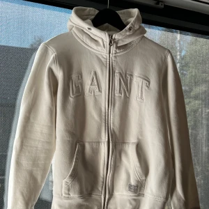 Vit hoodie från GANT - Säljer en stilren vit hoodie från GANT med dragkedja och stor logga på bröstet. Tröjan har en praktisk huva och fickor framtill. Perfekt för en avslappnad men stilren look. Skick: nästan oandvänd