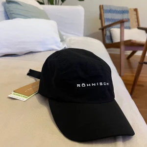 NY Svart keps från Röhnisch - Snygg svart keps från Röhnisch med justerbar rem baktill. Perfekt för en sportig look eller en avslappnad dag ute. Märkesloggan är broderad framtill i vitt.