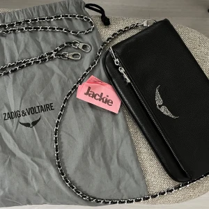 Svart axelväska Zadig & Voltaire skinn - Snygg svart axelväska från Zadig & Voltaire i äkta skinn med silverfärgad kedjerem och dragkedjedetaljer. Väskan har ett stilrent vingemblem framtill och praktisk dragkedja både fram och bak. Perfekt storlek för det viktigaste och ger en cool vibe.