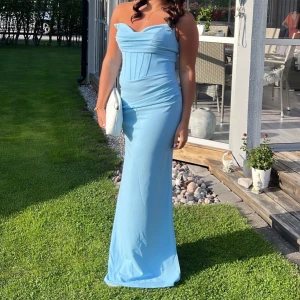 Ljusblå långklänning med snörning - Supersnygg ljusblå långklänning med axelbandslös design och figurnära passform. Klänningen har snygga draperingar upptill, markerad midja och snörning i ryggen för extra detalj. Perfekt för bal eller student!              Endast använd 1 gång