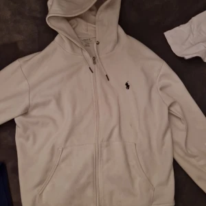 Polo Ralph Lauren hoodies med dragkedja - Två klassiska hoodies från Polo Ralph Lauren, en i vitt och en i svart. Båda har huva med snörning, dragkedja framtill och känguruficka. Ikoniska Polo-loggan broderad på bröstet. Perfekt basic till garderoben. 300 kr st 