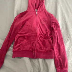 Rosa velour hoodie från Juicy Couture - Juicy Couture hoodie i härligt rosa velour med dragkedja och huva. Klassisk logga på bröstet och snygga fickor framtill. Väldigt sliten! Perfekt för dig som vill ha den ikoniska Y2K-looken och mjukaste känslan.