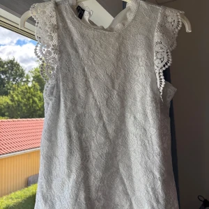 Vit spetsblus med volangärm - Superfin vit blus från pieces med vackra spetsdetaljer och volang vid ärmarna. Blusen är ärmlös och har ett romantiskt mönster i spets över hela plagget. Perfekt för dig som gillar en söt och stilren look!