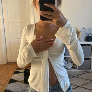 Vit omlottopp med knyt från Gina Tricot - Supersnygg vit omlottopp från Gina Tricot i mjuk bomullsmix. Långärmad modell med v-ringning och långa knytband framtill som ger en cool och trendig look. Perfekt att styla med jeans eller kjol för en fräsch vibe.