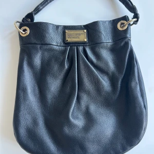 Svart väska Marc by Marc Jacobs - Snygg svart väska från Marc by Marc Jacobs i skinn med guldfärgade metalldetaljer. Perfekt väska för både skola och jobb, ett långt band ingår! Väskan är i bra skick, bara en guldig metall sak som fattas från brickan och lite sprucket i skinnet i handtaget, se bild 2 och 3. Pris kan diskuteras i snabb affär😊
