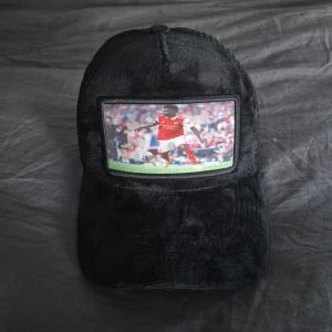 Svart meshkeps med fotbollstryck - Cool svart keps med mjuk sammet framtill och mesh baktill för extra ventilation. På framsidan sitter en patch med fotbollsspelaren bukayo saka på. Justerbar snapback baktill så den passar de flesta. Perfekt för dig som gillar sportig streetstyle.