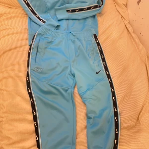 Nike ljusblå mjukisbyxor S - Nike mjukisbyxor i ljusblått med svarta och vita loggor längs sidorna. Byxorna har resår i midjan, sidofickor och är gjorda i mjukt syntetmaterial. Perfekta för chill eller träning, snygg sportig look.