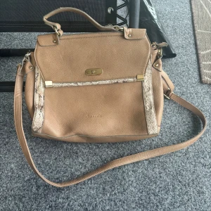 Beige axelväska från Tamaris - Snygg beige axelväska från Tamaris i skinnimitation med guldfärgade metalldetaljer och ormmönstrade inslag framtill. Väskan har både handtag och lång axelrem samt dragkedjeficka på baksidan. Perfekt för dig som vill ha en stilren och trendig väska.