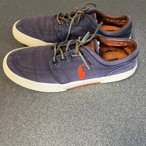 Lila sneakers från Polo Ralph Lauren - Snygga lila sneakers från Polo Ralph Lauren med klassisk orange logga på sidan. Skorna har vita sulor, rund tå och snörning. Ovandelen är i canvas och insidan har bruna detaljer. Perfekta för en chill och stilren look.