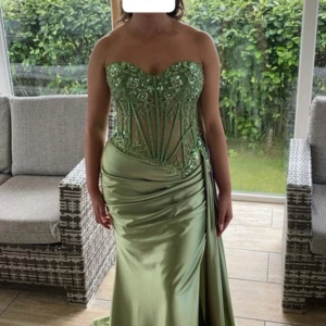 Grön balklänning från Ladivine - Snygg ljusgrön långklänning med axelbandslös, figurnära passform. Överdelen har transparent mesh med broderade blommor och pärlor, samt markerad midja. Kjolen är i glansigt satinliknande material med drapering och släp. Perfekt för bal eller student!