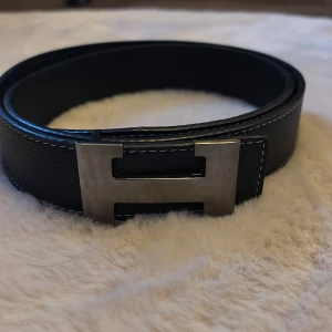 Svart skärp i skinn med vit söm - Säljer ett stilrent svart bälte i skinn med tydliga vita sömmar längs kanterna. Bältet är klassiskt rakt och passar perfekt till jeans eller kostymbyxor. Enkel design utan spänne på bilderna, vilket ger en clean look. 