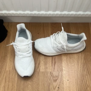 Vita Adidas Ultraboost sneakers - Säljer ett par helvita Adidas Ultraboost sneakers med stickad ovandel och klassiska tre ränder på sidan. Skorna har snörning, platt sula och bekväm Boost-dämpning. Perfekta för dig som gillar stilrena och sportiga sneakers.