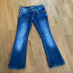 Miss Me blå bootcut jeans med paljetter - Snygga blå jeans från Miss Me med bootcut passform och coola paljettbroderier på bakfickorna thight uppe vid midjan. Jeansen har slitna detaljer, vita kontrastsömmar och dekorativa nitar. Perfekta för dig som vill sticka ut med lite extra bling.