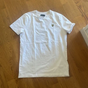 Vit t-shirt från Lyle & Scott Junior - Vit t-shirt från Lyle & Scott Junior med klassisk passform och rund hals. T-shirten har ett broderat gult och svart logomärke på bröstet och är tillverkad i mjuk bomull. Perfekt basic-plagg med stilren look.