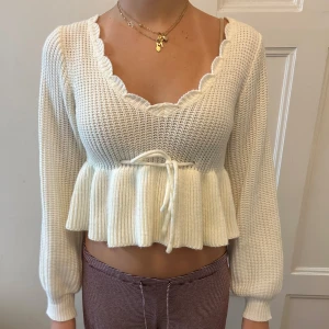 Vit stickad tröja med volang och knyt - Supersöt vit stickad tröja med volangkant nertill och scallop-detaljer vid halsringningen. Tröjan har lång ärm, croppad passform och knytband i midjan för extra söt look. Perfekt för lager-på-lager under hösten eller våren.