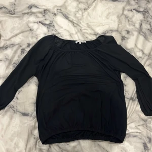 Svart blus från Flash Woman - En svart blus från Flash Woman med rundad halsringning och långa ärmar. Modellen är lite lösare och har resår i nederkanten för en snygg passform. Perfekt att styla med jeans eller kjol för en clean look.