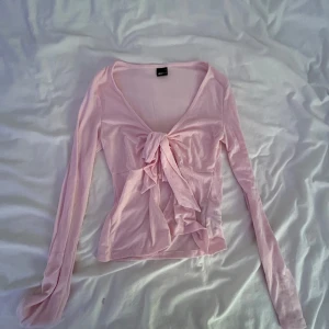 Ljusrosa omlottopp från Gina Tricot - Supersöt ljusrosa omlottopp från Gina Tricot med lång ärm och knyt framtill. Toppens material är mjukt och stretchigt, perfekt för en trendig och feminin look. Djup v-ringning och croppad passform gör den extra stylish.