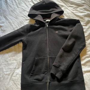 Svart hoodie från GANT med dragkedja - Svart hoodie från GANT med broderad logga på bröstet. Tröjan har huva, hel dragkedja och två stora fickor framtill. Tillverkad i mjuk bomullsmix, perfekt för chill dagar. Klassisk och stilren design som funkar till det mesta. 158-164 cm