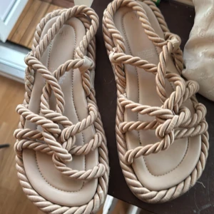 Beiga sandaler med repdetaljer - Snygga sandaler i beige med unika repdetaljer som ger en cool och trendig look. Sulan är platt och mjuk, och remmarna är tvinnade för en bohemisk känsla. Perfekta för varma dagar och sommaroutfits.