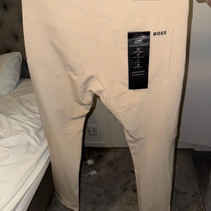 Beige byxor från BOSS, storlek 48 - Snygga beige byxor från BOSS i stretchigt material med raka ben och diskret logga på bakfickan. Byxorna har dragkedja i fickan och är perfekta för en clean och stilren look. Passar dig som gillar klassisk men modern stil.
