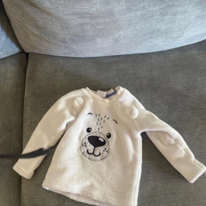 Vit fleece tröja med björnansikte baby  - Storlek 56 — Supermjuk vit fleece tröja med ett gulligt björnansikte broderat framtill och små öron på axlarna. Perfekt för mysiga dagar och har långa ärmar. Passar dig som gillar söta detaljer och vill ha något riktigt bekvämt.