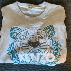 Grå Kenzo sweatshirt med tigertryck - Säljer en grå sweatshirt från Kenzo i storlek XS med det ikoniska tigerbroderiet i silver, blått och svart på framsidan. Tröjan har rund hals, långa ärmar och är gjord i mjuk bomull. Perfekt för dig som gillar statement-plagg och coola designer.