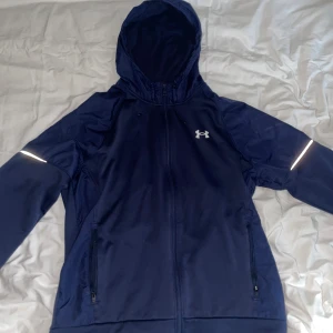 Marinblå vindjacka Under Armour - Snygg marinblå vindjacka från Under Armour med huva, hel dragkedja och reflekterande detaljer på ärmarna. Jackan har två fickor med dragkedja och logga på bröstet samt ryggen. Materialet är polyester och jackan är perfekt för träning utomhus.
