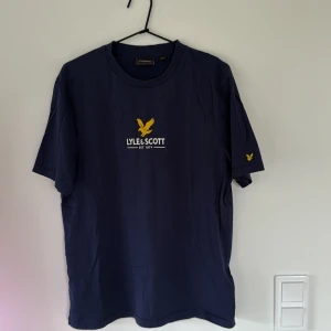Marinblå t-shirt från Lyle & Scott - Snygg marinblå t-shirt från Lyle & Scott med gul logga och text på bröstet. Klassisk rund hals och korta ärmar. Mjuk bomullskvalitet som känns skön mot huden. Perfekt för dig som gillar stilrena och sportiga plagg.