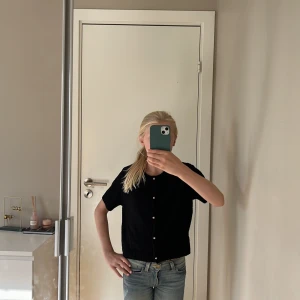 Svart stickad kofta från H&M - Svart stickad kofta från H&M med korta ärmar och rund hals. Koftan har snygga guldfärgade knappar framtill och ribbade muddar vid ärmslut och nederkant. Perfekt att slänga över en t-shirt för en chill look.