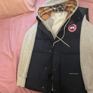 Mörkblå dunväst från Canada Goose - Snygg mörkblå dunväst från Canada Goose med klassisk logga på bröstet och diskret broderad text. Västen har stora fickor framtill, knappar och dragkedja. Perfekt för lager-på-lager och har en clean, urban vibe.