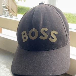 Svart keps från BOSS med tryck - Svart keps från BOSS med stort guldigt BOSS-tryck framtill. Klassisk böjd skärm och justerbar baksida. Insidan har unika guldmönster och BOSS-logga på etiketten. Perfekt accessoar för en cool och sportig look.