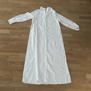 Vit Lucia långklänning med krage - En stilren vit Lucia klänning med lång ärm och klassisk krage. Lucia klänningen har rak passform och är tillverkad i ett lätt tyg som ger en luftig känsla. Perfekt för dig som gillar minimalistisk och clean stil.