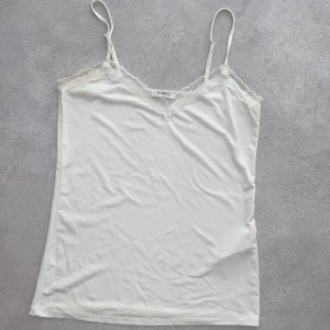 Vit linnetopp med spets från Pieces - Supersöt vit linnetopp från Pieces med tunna justerbara axelband och v-ringning dekorerad med spets. Toppens material är mjukt och följsamt, perfekt för lager-på-lager eller som statement-piece. Enkel och stilren design som är lätt att matcha.