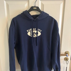 Mörkblå hoodie från Lee  - Snygg mörkblå hoodie från Lee med vitt Kansas City-tryck på bröstet. Tröjan har huva med snörning och är långärmad. Regular fit och tillverkad i mjuk bomull, perfekt för chill dagar eller när du vill ha en avslappnad look. Mindre i storlek