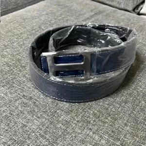 Mörkblå  skärp med H-spänne - Snyggt mörkblått skärp från med ett stort, silvrigt H-spänne i metall. Bältet har vita sömmar längs kanterna och är tillverkat i slätt skinn. Perfekt för att lyfta din outfit med en exklusiv touch.