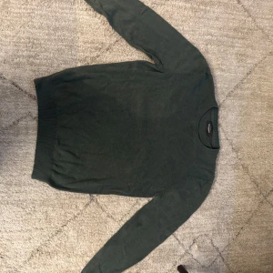 Mörkgrön stickad tröja från Dressmann - Snygg mörkgrön stickad tröja från Dressmann i storlek S. Tröjan har rund hals och långa ärmar med ribbade muddar vid hals, ärmslut och nederkant. Perfekt för dig som gillar enkel och stilren design.