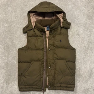 Olivgrön dunväst från Polo Ralph Lauren - Snygg olivgrön dunväst från Polo Ralph Lauren med avtagbar huva och dragkedja framtill. Västen har två stora fickor, diskret logga på bröstet och beige foder. Perfekt för lager-på-lager och chill höst- eller vinterstil.