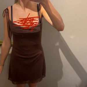 Brun kort klänning från Stradivarius - Supersnygg brun kort klänning från Stradivarius med tunna axelband som knyts på axlarna. Klänningen har en lätt figurnära passform och är tillverkad i ett mjukt syntetmaterial. Nederkanten har en diskret volang för extra vibe. 