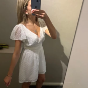 Vit kortklänning med prickigt mönster - Supersöt vit kortklänning med v-ringad hals och korta, luftiga ärmar. Klänningen har ett diskret prickigt mönster i tyget och ett markerat midjeband som ger en snygg siluett. Perfekt för sommarens alla tillfällen och riktigt fräsch stil.