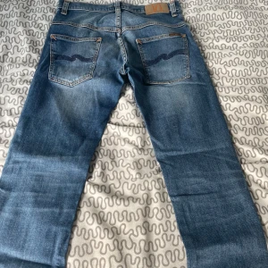 Blå jeans från Nudie Jeans Co, W30 L30 - Säljer ett par klassiska blå jeans från Nudie Jeans Co i storlek W30 L30. Jeansen har slitna detaljer framtill, orangea sömmar och Nudie-logga på bakfickorna. De är tillverkade i jeansmaterial och har en rak passform med normal midja.