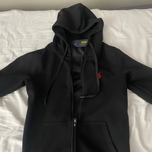 Svart hoodie från Polo Ralph Lauren - Svart hoodie från Polo Ralph Lauren med röd broderad logga på bröstet. Tröjan har huva med snörning, dragkedja framtill och två fickor. Materialet är mjuk bomull och passformen är normal. Perfekt för chill dagar och streetwear-stil.