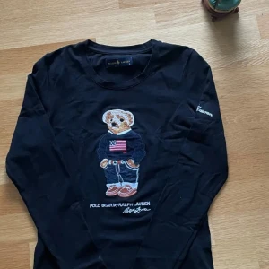 Svart långärmad tröja Polo Bear - Svart långärmad tröja från Ralph Lauren med ikonisk Polo Bear broderad framtill. Tröjan har rund hals och coolt broderi med björnen i jeans och tröja med amerikansk flagga. Mjuk bomullskvalitet och normal passform. Perfekt för dig som gillar street och klassisk stil.